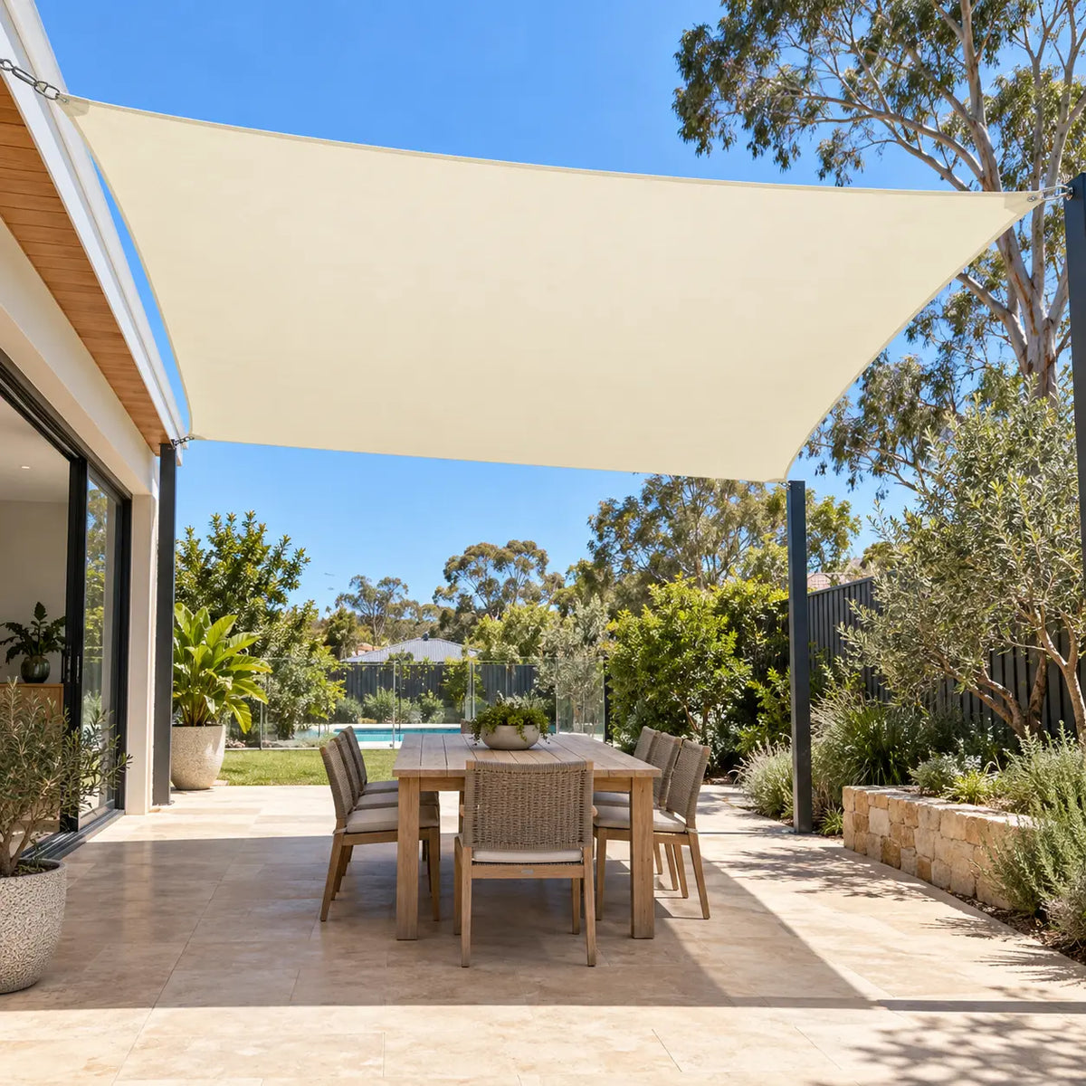 Cream shade sail rectangle over outdoor dining table with wicker chairs beside native garden and pool in Australian backyard