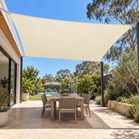 Cream shade sail rectangle over outdoor dining table with wicker chairs beside native garden and pool in Australian backyard