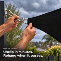 Unclipping a black shade sail snap hook from a wall pad eye before a storm